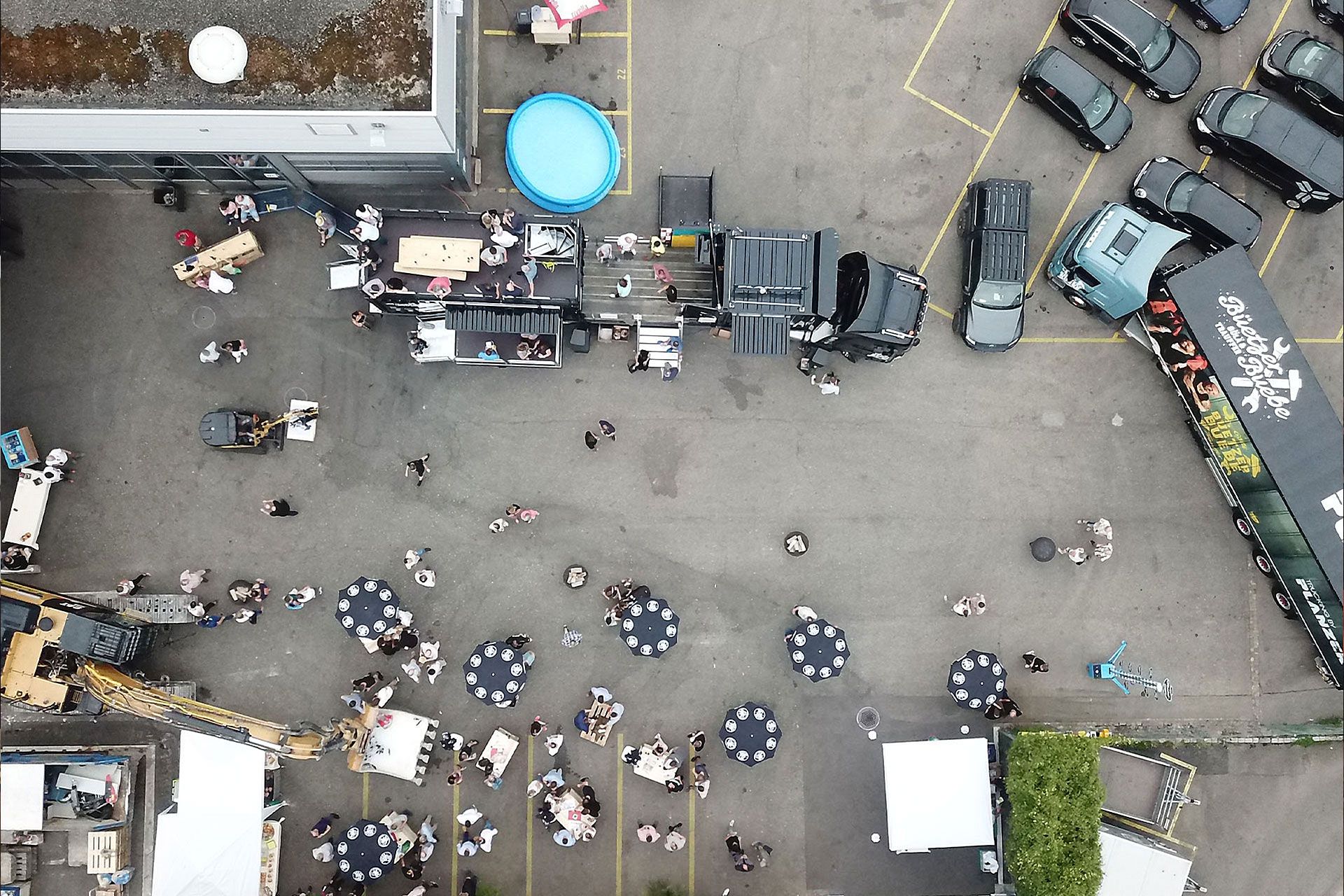 Luftaufnahme eines Outdoor-Events mit Menschen, die sich um den Event-Truck mit geöffneter Bühne und Dachterrasse versammeln, umgeben von Sonnenschirmen und parkenden Fahrzeugen.