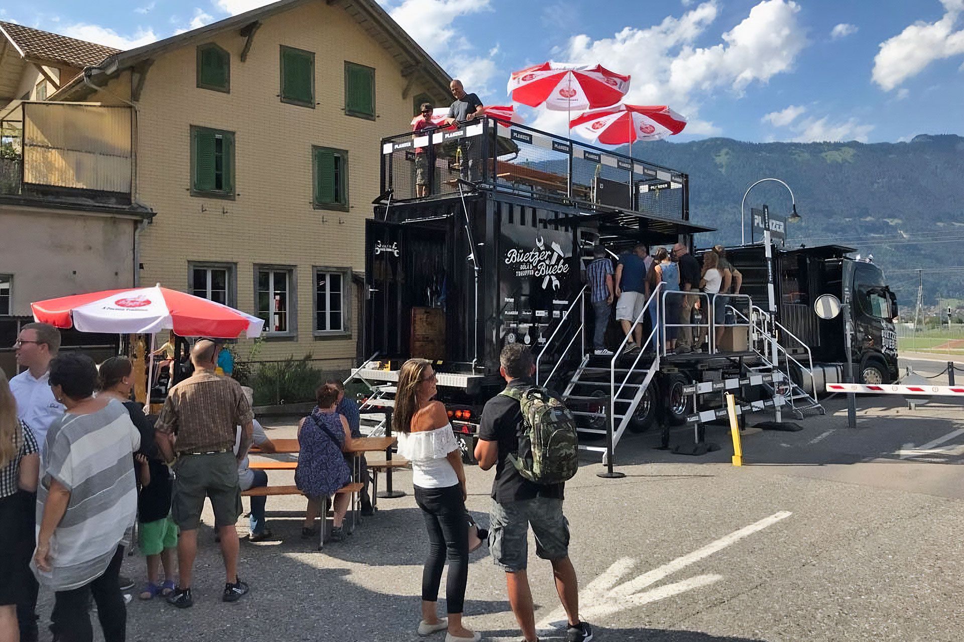 Besucher versammeln sich um den Planzer Event-Truck mit Dachterrasse und Sonnenschirmen bei einem öffentlichen Event an einem sonnigen Tag.
