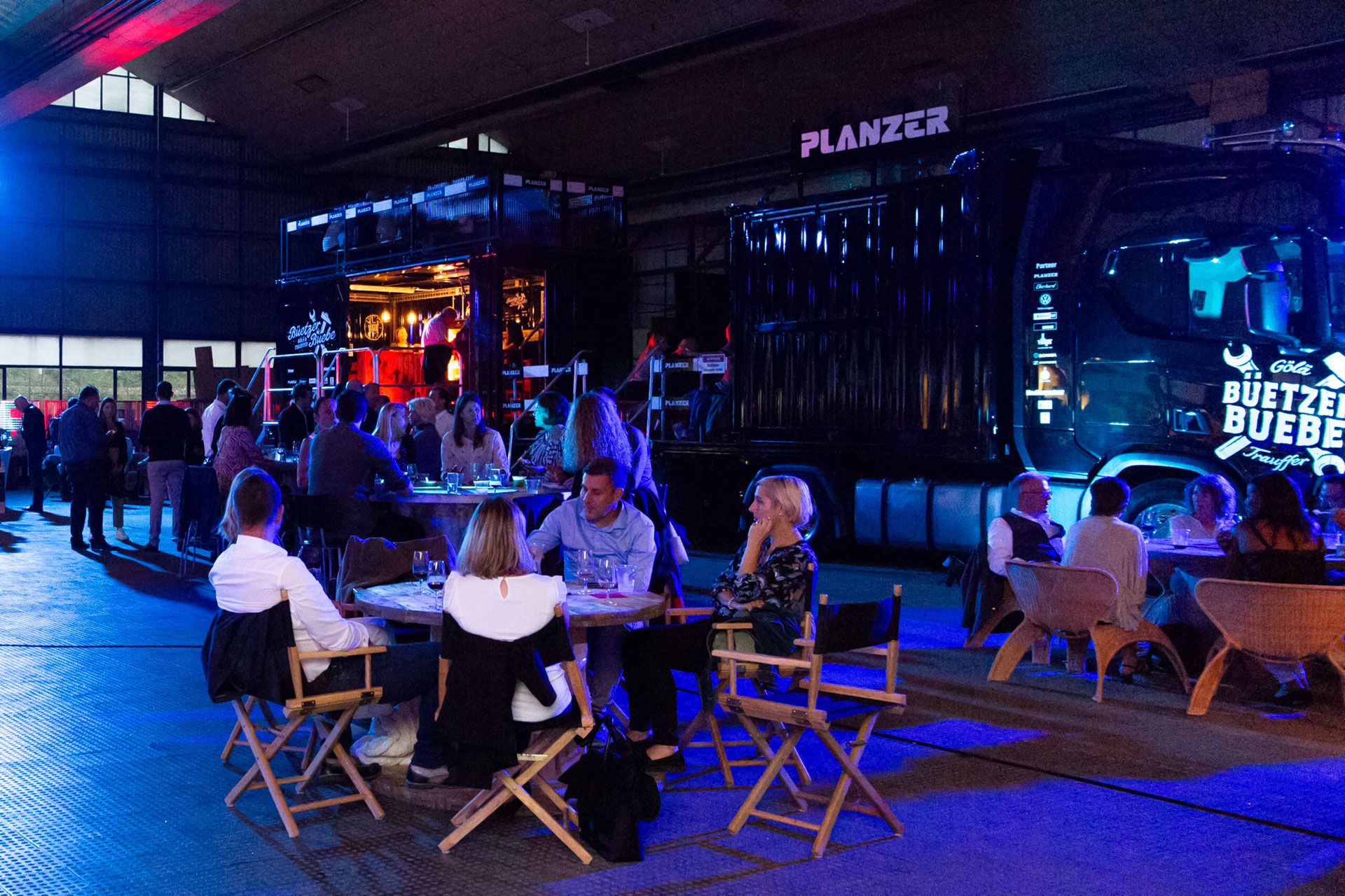 Besucher sitzen und unterhalten sich bei einem Abendevent vor einem beleuchteten Planzer Event-Truck in einer Industrieumgebung.